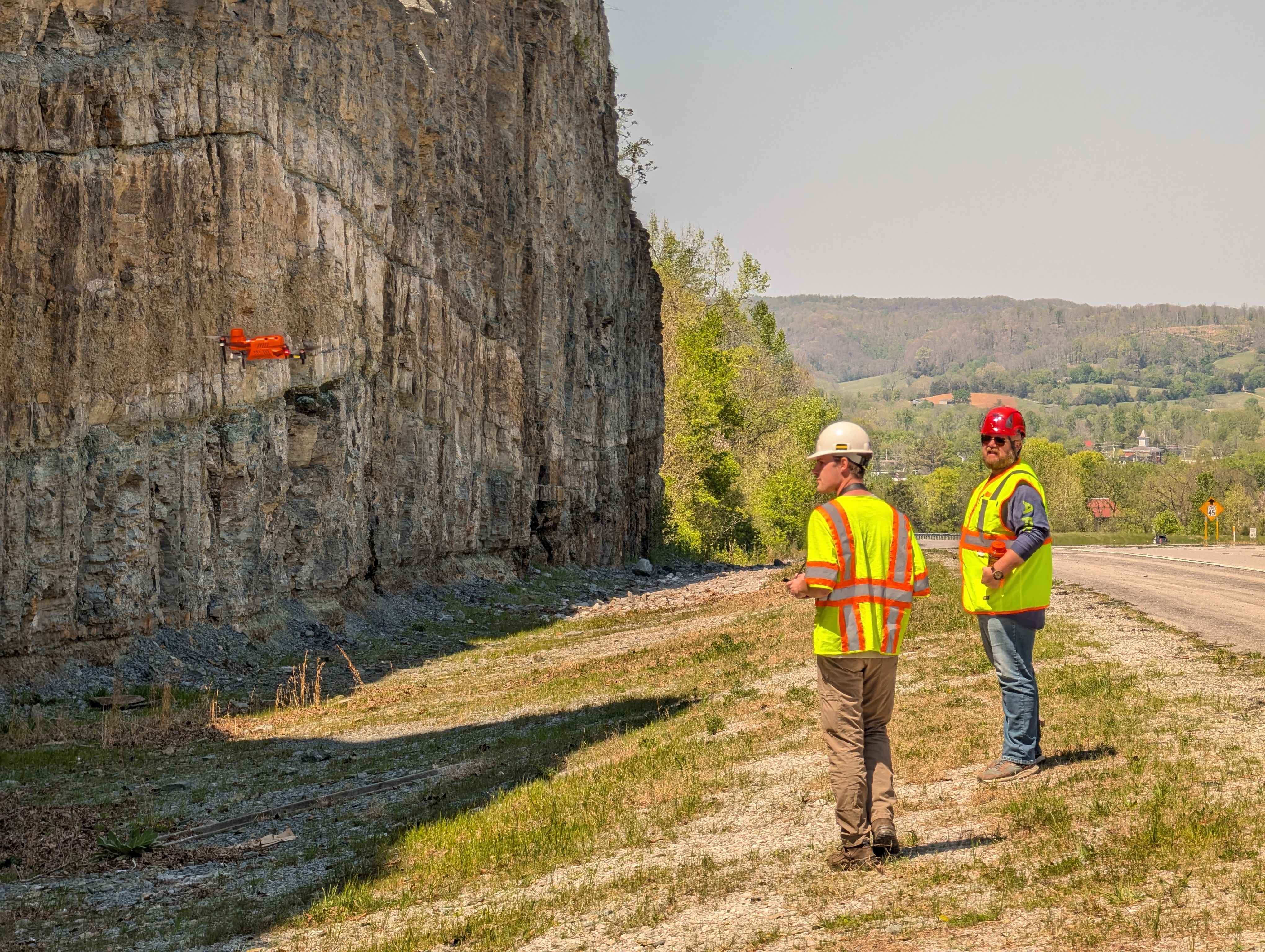 Two researchers wearing safety equipment monitor an orange UAV flying beside a large rock roadcut in Celina, Tennessee