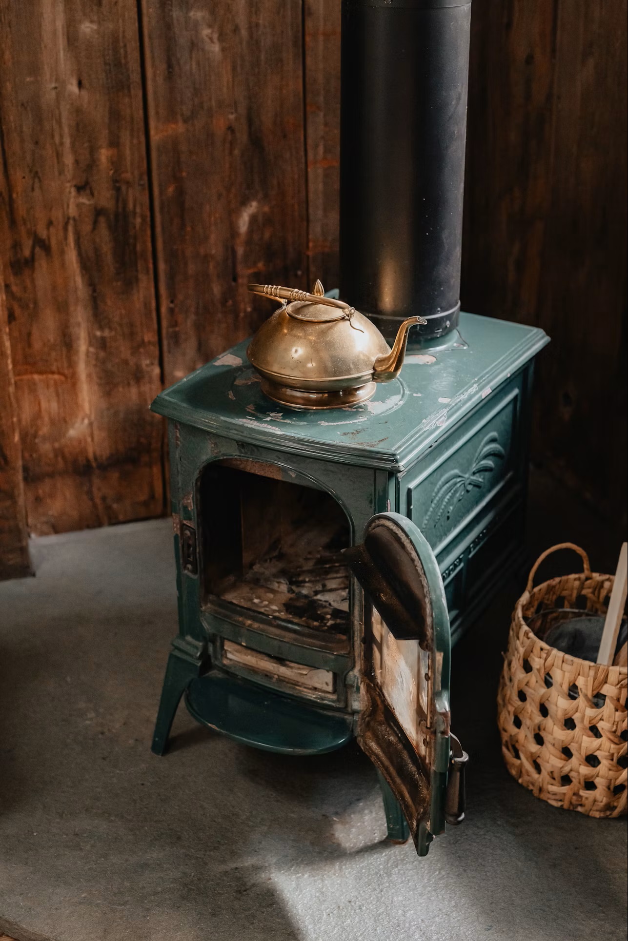 A green residential wood stove with its door open and a metal kettle resting on top
